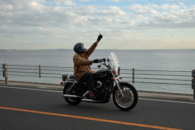 Motorcyclist riding along a coastal road with one hand raised toward the cloudy sky.